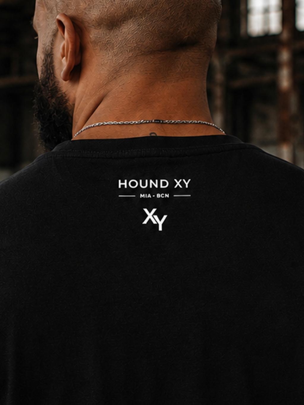A man viewed from behind wearing a black crewneck t-shirt with a white 'HOUND XY' logo printed on the upper back, set against a dark, b