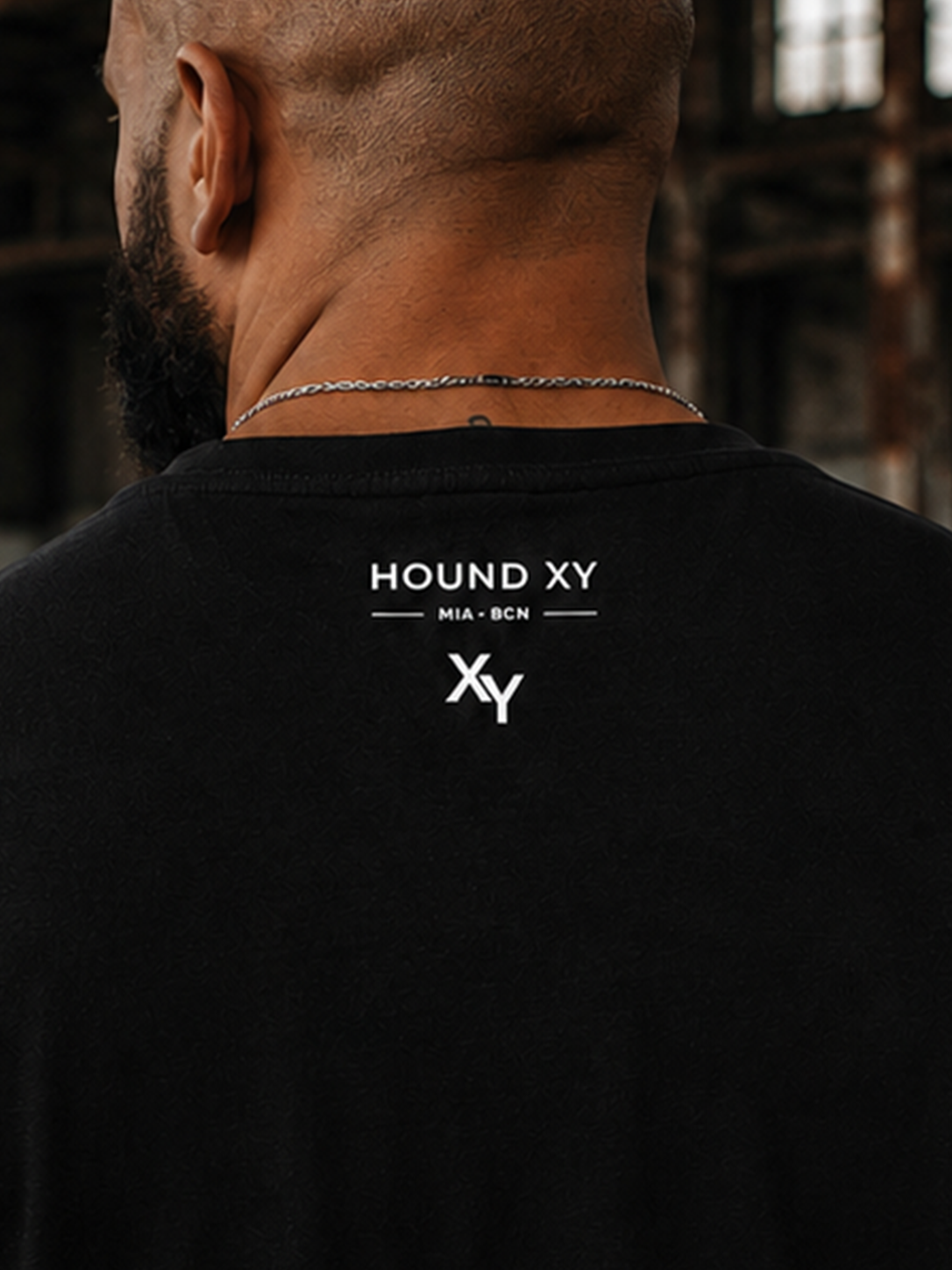 A man viewed from behind wearing a black crewneck t-shirt with a white 'HOUND XY' logo printed on the upper back, set against a dark, b